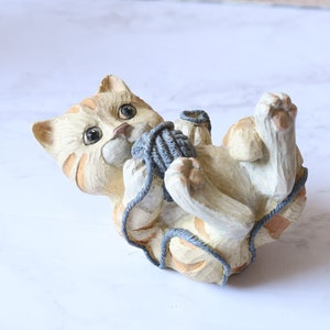 May include: A ceramic figurine of a tan and orange tabby cat playing with a ball of blue yarn. The cat is lying on its back with its paws in the air.