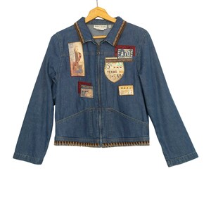 May include: A blue denim jacket with a zipper closure and a brown trim around the collar and bottom hem. The jacket has several patches sewn on the front, including a patch with the text "FA70S COLORADO" and another patch with the text "JS TEXAS 9-SN1".