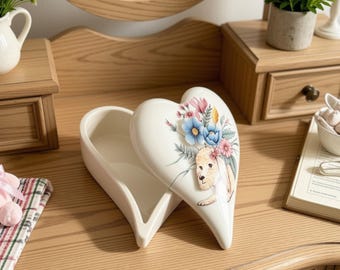 May include: A white heart-shaped ceramic trinket box with a lid featuring a painted illustration of a light brown dog surrounded by blue, pink, and yellow flowers. The box is open, revealing a smooth, hollow interior for storing small items.