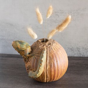 May include: A brown ceramic vase with a textured surface and a sculpted octopus design. The vase is filled with dried beige grasses.