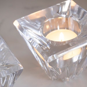 May include: Two clear glass candle holders with a square base and a beveled design. Each holder has a small, round opening at the top for a tea light candle.