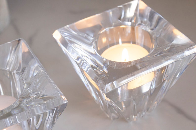 May include: Two clear glass candle holders with a square base and a beveled design. Each holder has a small, round opening at the top for a tea light candle.