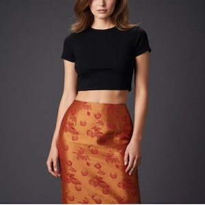 May include: A black short-sleeved crop top paired with a high-waisted midi skirt in a metallic copper-orange hue. The skirt features a repeating woven pattern of circular floral motifs and stylized bird figures in a darker shade of red-orange.