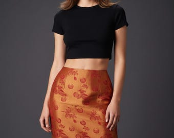 May include: A black short-sleeved crop top paired with a high-waisted midi skirt in a metallic copper-orange hue. The skirt features a repeating woven pattern of circular floral motifs and stylized bird figures in a darker shade of red-orange.
