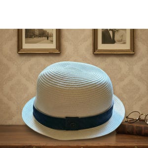 May include: A light-colored fedora hat with a dark blue band. The hat has a round crown and a brim. The band is secured with a small button. The hat is made of a woven material.
