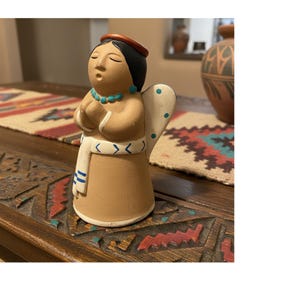 May include: A handcrafted ceramic angel figurine with closed eyes and folded hands. The angel wears a turquoise necklace, a red headband, and a white sash with blue stripes. The wings are white with blue dots.