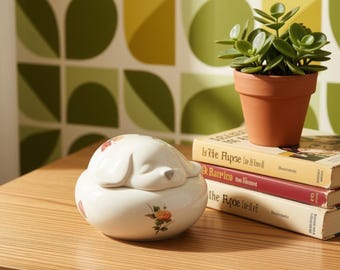 May include: A white ceramic trinket box with a lid shaped like a sleeping dog, featuring small painted orange rose motifs. The box sits beside a terracotta pot containing a green succulent plant and a stack of three books with visible text.