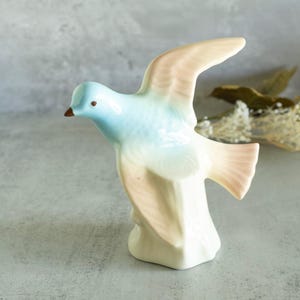 May include: A ceramic bird figurine with a light blue and pink glaze. The bird is perched on a white pedestal with a pink base. The figurine is a vintage style.