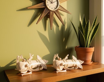 May include: Two cream-colored woven lobster figurines with gold-toned accents and spherical eyes sit on a wooden shelf. Above them, a gold starburst wall clock hangs on a light green wall next to a potted snake plant in a terracotta pot.
