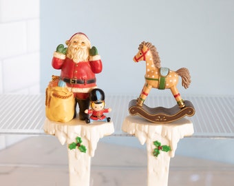 May include: Two white ceramic Christmas tree toppers. One features Santa Claus holding a bag of gifts and a toy soldier. The other features a rocking horse with brown and white spots.