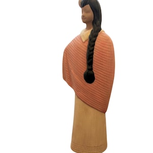 May include: A ceramic figurine of a Native American woman. The figure has long black hair in a braid, a peach-colored shawl, and a light tan dress. The figurine is standing and has a serene expression.