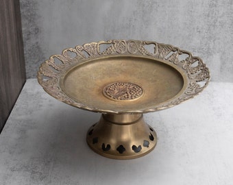 May include: A vintage brass pedestal dish with an ornate, openwork rim featuring grape and leaf designs. The center of the dish has a raised medallion with a grape cluster. The pedestal base has decorative cutouts.
