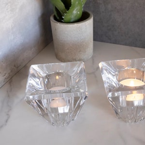 May include: Two clear glass candle holders with a square base and a pyramid-shaped design. The candle holders are on a white marble surface and each has a small white tea light candle inside.