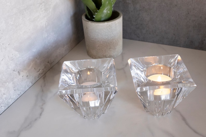 May include: Two clear glass candle holders with a square base and a pyramid-shaped design. The candle holders are on a white marble surface and each has a small white tea light candle inside.