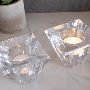 May include: Two clear glass candle holders with a square base and a pyramid-shaped top. Each holder has a small, round candle inside.