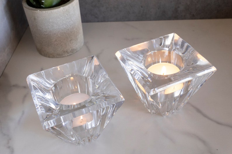May include: Two clear glass candle holders with a square base and a pyramid-shaped top. Each holder has a small, round candle inside.