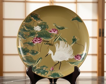 May include: A round decorative plate with a gold-toned background featuring a white egret standing among green lotus leaves and pink and white lotus flowers. The plate is supported by a dark brown wooden stand, showcasing detailed botanical art.