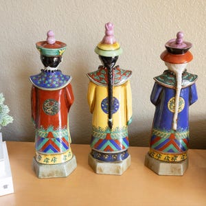 May include: Three ceramic figurines of people wearing traditional Chinese clothing. The figurines are painted in bright colors, including red, blue, yellow, and green. They are standing on a wooden surface.