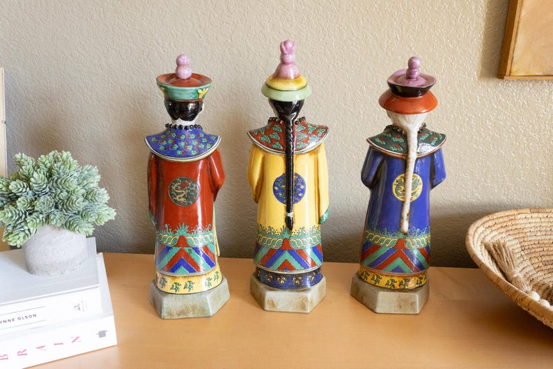 May include: Three ceramic figurines of people wearing traditional Chinese clothing. The figurines are painted in bright colors, including red, blue, yellow, and green. They are standing on a wooden surface.