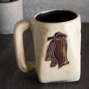 May include: A beige ceramic mug with a brown handle. The mug has a stylized design of a brown, red, and purple flower on the front.