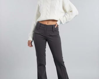 May include: A long-sleeve white cable knit sweater paired with charcoal gray trousers. The trousers feature a mid-rise waist, two front button-flap pockets, a double-button closure, and a straight-leg cut with a pressed crease down the center.