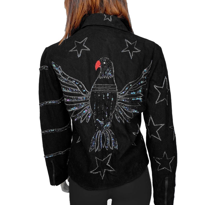K&ouml;nnte beinhalten: Schwarze Wildlederjacke mit einem Pailletten-Adler-Design auf der R&uuml;ckseite. Der Adler hat einen roten Schnabel und ist von silbernen Sternen umgeben.