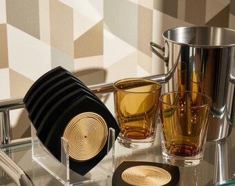 May include: A set of black square coasters with circular beige spiral inlays, stored in a clear acrylic holder. Two amber-colored glass tumblers and a stainless steel ice bucket are positioned nearby on a glass surface with a chrome frame.
