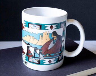 May include: White ceramic mug with a Southwestern design. The mug features a turquoise and brown geometric border and a scene depicting figures in traditional clothing against a desert landscape. The word "DESERT" is printed on the mug.