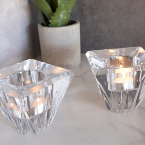 May include: Two clear glass candle holders with a geometric design. Each holder has a square base and a pyramid-shaped top. The holders are empty and ready to be filled with tea lights.