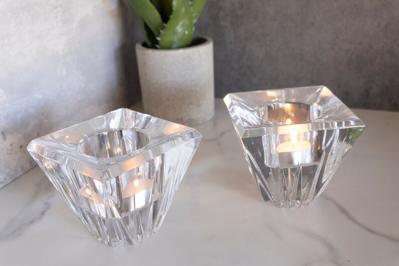May include: Two clear glass candle holders with a geometric design. Each holder has a square base and a pyramid-shaped top. The holders are empty and ready to be filled with tea lights.