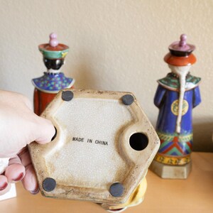 May include: A ceramic figurine of a person wearing a colorful robe and hat. The figurine is standing on a hexagonal base with the words "Made in China" printed on the bottom.