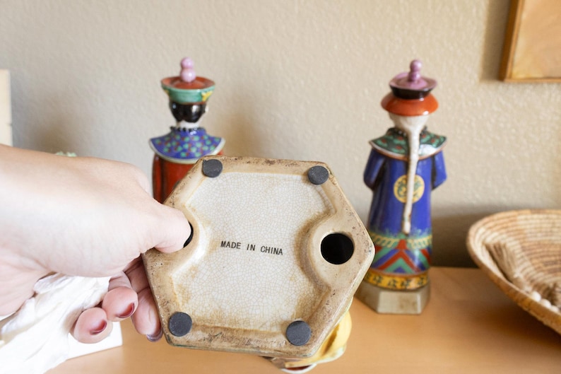 May include: A ceramic figurine of a person wearing a colorful robe and hat. The figurine is standing on a hexagonal base with the words "Made in China" printed on the bottom.