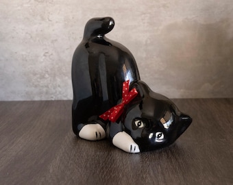 May include: Black ceramic cat figurine with a red polka dot bow. The cat is lying on its back with its paws in the air.