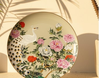 May include: A round decorative plate with a cream-colored background featuring a white peacock with a detailed tail, surrounded by vibrant pink, white, and red peony flowers with green leaves and a thin gold rim along the edge of the plate.