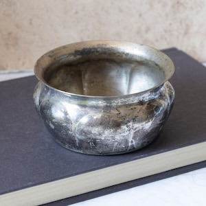 May include: A silver bowl with a slightly worn finish. The bowl has a rounded shape with a slightly flared rim. The bowl is sitting on a dark gray book.