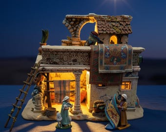 May include: A miniature stone-colored building with a tiled roof, a wooden ladder, and a hanging patterned rug. Two figures stand in front, one holding a blue cloth. The structure is illuminated from within, casting a warm glow against a dark background.