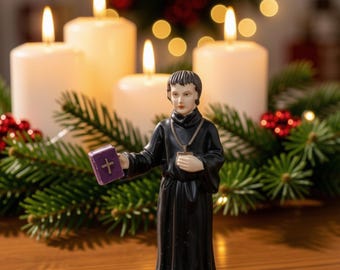 May include: A figurine of a person wearing a black robe and a cross necklace, holding a small purple book with a gold cross on the cover. The figure stands in front of four white pillar candles and green pine branches with red berries.