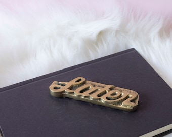 May include: A gold-colored metal word plaque that reads "later" on a dark gray notebook. The notebook is on a white fluffy surface.