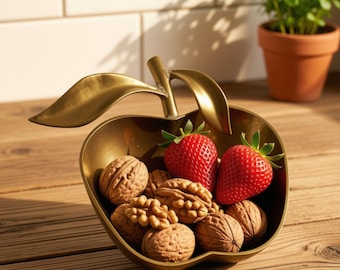 May include: A metallic gold apple-shaped bowl featuring a stem and two leaves, filled with several brown walnuts and two bright red strawberries with green stems. The bowl has a smooth, reflective finish and a curved, open-top design.