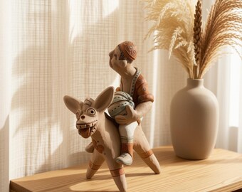 May include: A terracotta-colored ceramic figurine depicting a person riding a stylized four-legged animal. The person wears a patterned cap and shirt, holding a round, striped object. The animal has large ears, a wide mouth, and painted details.