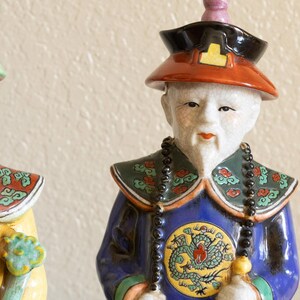 May include: Two porcelain figurines of people wearing traditional Chinese clothing. The figurine on the left is wearing a yellow and green robe with a black beaded necklace. The figurine on the right is wearing a blue and green robe with a black beaded necklace. Both figurines have painted facial features and elaborate headdresses.
