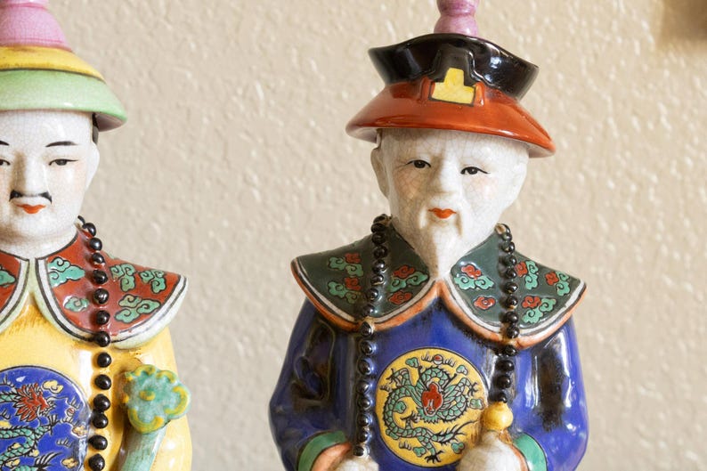 May include: Two porcelain figurines of people wearing traditional Chinese clothing. The figurine on the left is wearing a yellow and green robe with a black beaded necklace. The figurine on the right is wearing a blue and green robe with a black beaded necklace. Both figurines have painted facial features and elaborate headdresses.