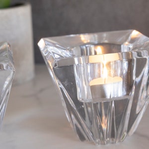 May include: Two clear glass candle holders with a geometric design. The candle holders are filled with tea lights that are lit.
