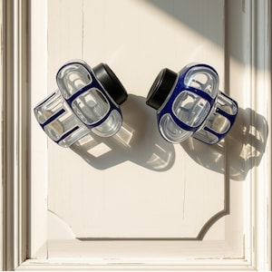 May include: Two cabinet knobs featuring a clear glass body with a deep blue grid pattern and a solid black circular base. The knobs are mounted on a light-colored paneled surface, casting distinct shadows under bright, directional lighting.