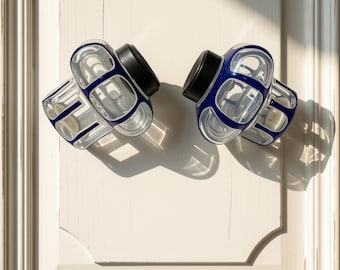 May include: Two cabinet knobs featuring a clear glass body with a deep blue grid pattern and a solid black circular base. The knobs are mounted on a light-colored paneled surface, casting distinct shadows under bright, directional lighting.
