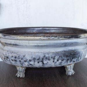 May include: A distressed, antique-style metal bowl with a dark interior and light gray exterior. The bowl has two handles and rests on three claw feet. The exterior has a textured, aged appearance, suggesting it is vintage.