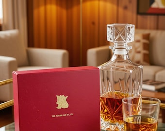 May include: A rectangular red box with a gold embossed logo and the text ACE PLAYING CARD CO., LTD. next to a clear glass decanter with a geometric stopper and a matching glass, both containing an amber liquid, on a gold metal tray.