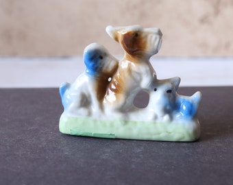 Peut inclure: Une figurine en porcelaine de trois chiens. Le plus grand chien est brun et blanc avec des accents bleus. Les deux plus petits chiens sont blancs avec des accents bleus. Les chiens sont debout sur une base verte.
