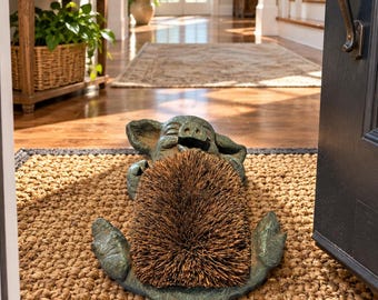 May include: A green, cast-metal pig-shaped boot scraper featuring a textured, brown, stiff-bristled brush insert on its back. The pig is depicted in a lying position with its head resting on its front hooves, positioned on a woven brown doormat.