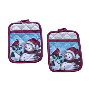 May include: A pair of red and white quilted pot holders with a snowman and cardinal design. The pot holders feature the text "Laura Ingalls Wilder".
